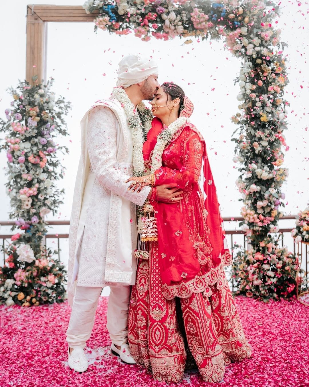 Bride and groom sharing a kiss under a floral arch, rose petals falling